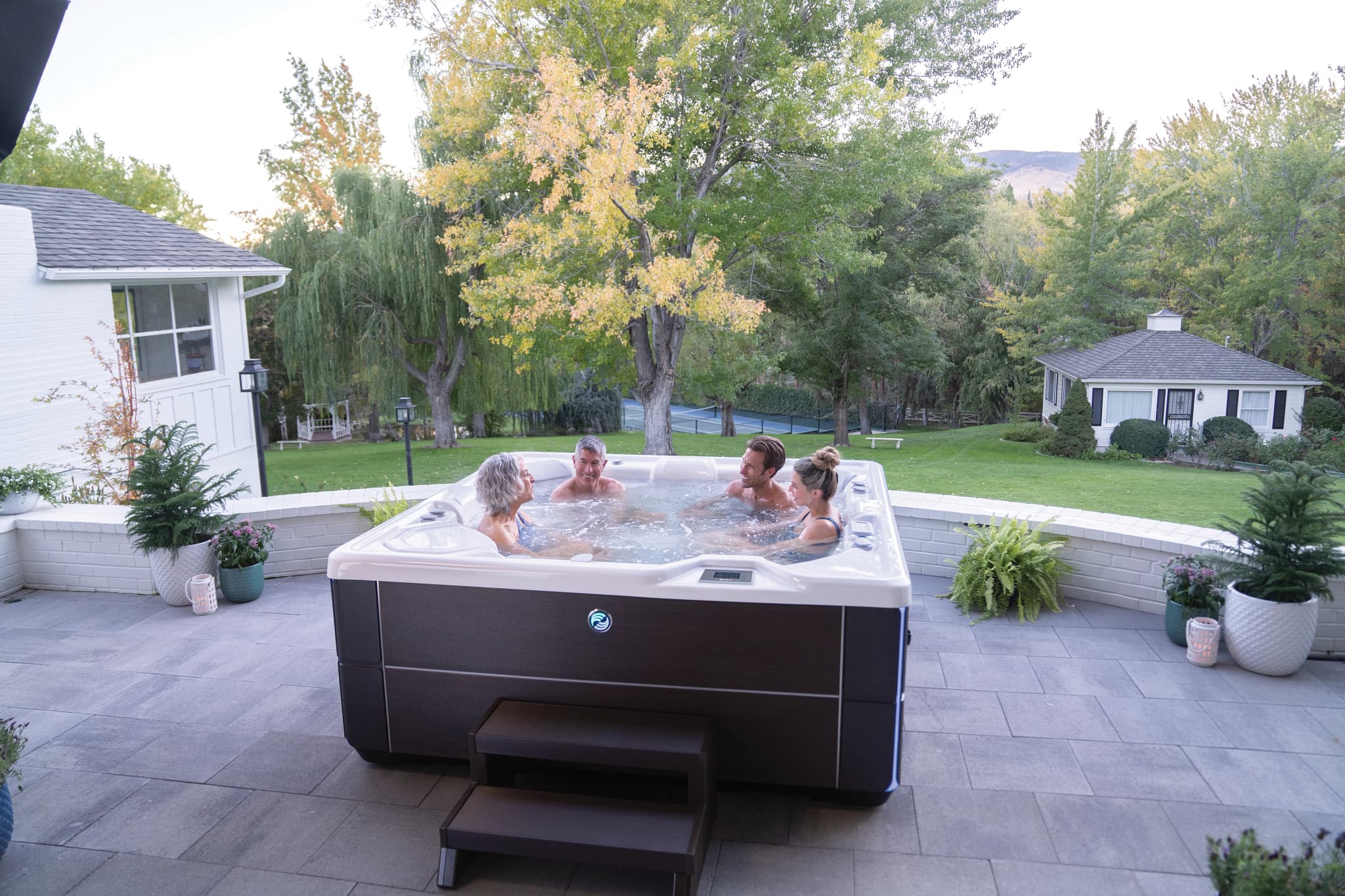 Four people talking in a Hot Spring Highlife Vanguard spa on a brick patio at dusk
